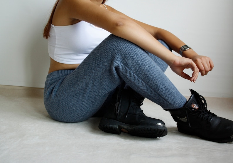 Close seated side view of denim blue knitted pants with raised cable detailing and chunky black footwear.