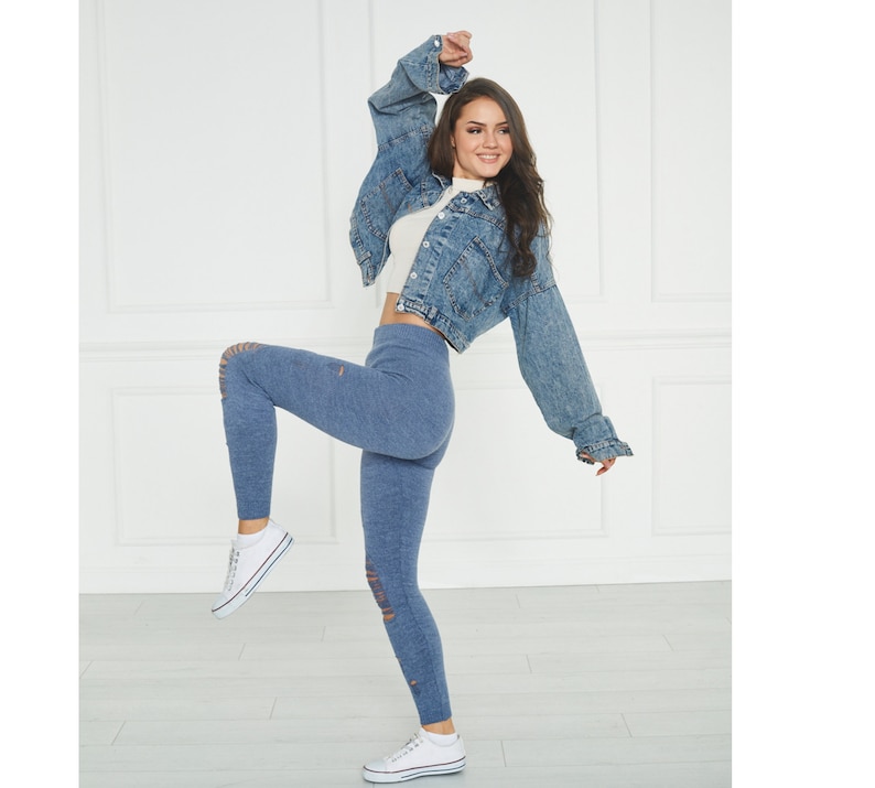 Puede incluir: Una chaqueta corta de mezclilla azul con cierre de botones y mangas largas, combinada con leggings de punto azul de talle alto con detalles desgastados en las rodillas, zapatillas de lona blancas con suela de goma y un top corto acanalado.