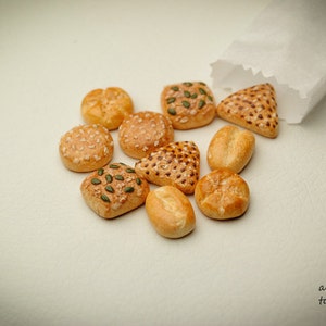 Könnte beinhalten: Eine Auswahl an Miniatur-Brötchen, darunter einfache, Sesam- und Mohnbrötchen, werden auf einer weißen Oberfläche gezeigt. Die Brötchen sind in einem Halbkreis angeordnet, wobei ein weißer Papierbeutel im oberen rechten Eck des Bildes teilweise sichtbar ist.