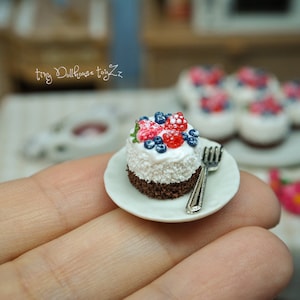 Könnte beinhalten: Ein Miniatur-Schokoladenkuchen mit weißem Frosting und roten und blauen Beeren darauf, der auf einem weißen Teller mit einer silbernen Gabel liegt. Der Kuchen liegt auf einer Hand.