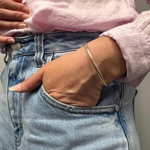 May include: A thin, polished gold-tone metal bangle bracelet with a smooth, rounded surface. The bracelet is worn on the wrist, featuring a simple, continuous circular design that reflects light across its metallic finish, creating a subtle shine.