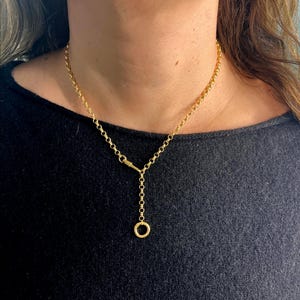 May include: A gold-colored necklace with a delicate chain and a distinctive Y-shaped design. The necklace features a small, round pendant at the end of the Y. The chain is composed of small, circular links.