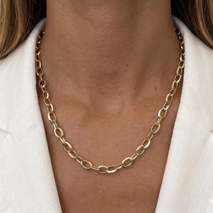 May include: A gold chain necklace featuring oval links. The necklace is displayed against a white blazer. The chain is a striking accessory, reflecting light and adding a touch of elegance. The necklace is a classic piece of jewellery.