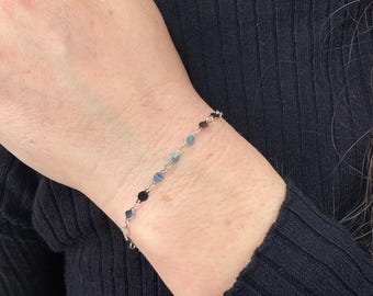 May include: A delicate silver chain bracelet featuring small, round, translucent blue beads spaced evenly along the metal links. The bracelet rests on a wrist, showcasing a simple design with alternating metallic silver chain segments and smooth, light blue circular stones.