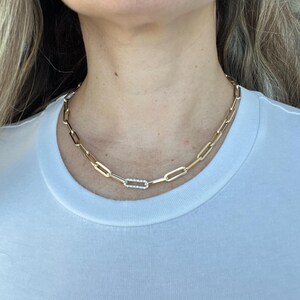 May include: A gold-toned paperclip chain necklace featuring a single rectangular link embellished with small clear stones. The necklace rests against the neck, showcasing a series of elongated, interlocking metal links in a polished finish.
