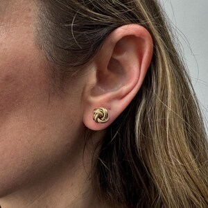 May include: Close-up of a person's ear with a gold-colored knot stud earring. The earring is a small, round design with a textured surface. The person has light skin and long, light brown hair.