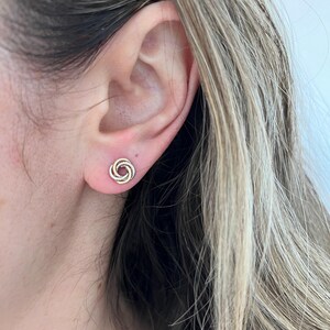 May include: A stud earring featuring a tri-color design with intertwined circular loops in yellow, rose, and white metal tones. The small, compact knot-shaped earring sits securely against the earlobe, showcasing a polished metallic finish.
