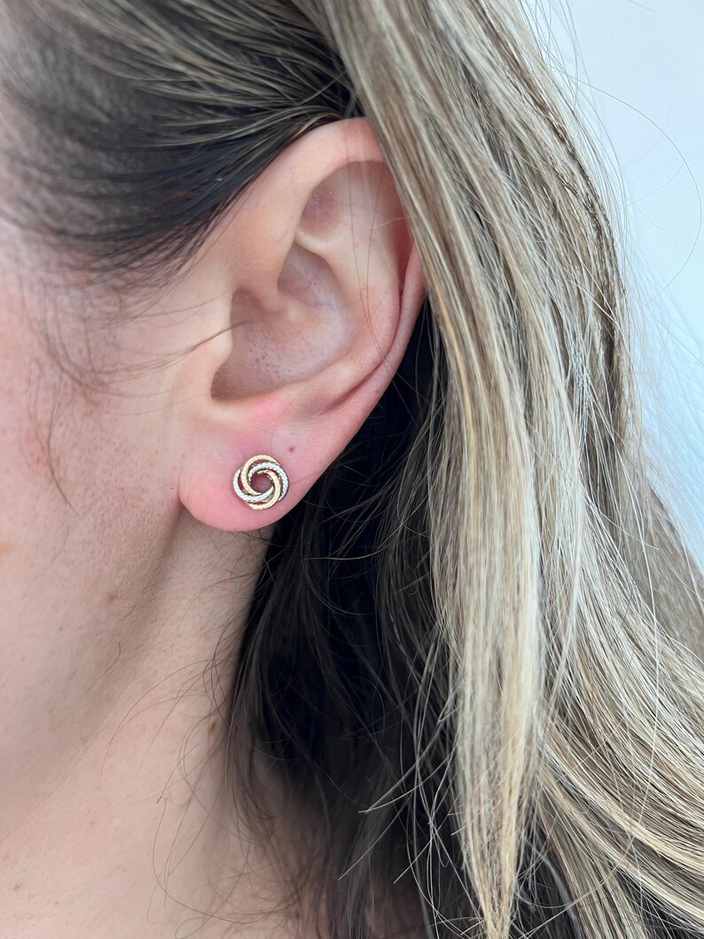 May include: A stud earring featuring a tri-color design with intertwined circular loops in yellow, rose, and white metal tones. The small, compact knot-shaped earring sits securely against the earlobe, showcasing a polished metallic finish.