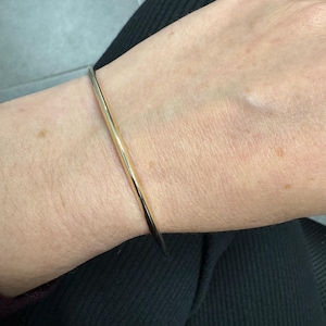 May include: A thin, polished gold-tone metal bangle bracelet with a smooth, rounded surface. The bracelet features a simple, continuous circular design that rests comfortably around the wrist, showcasing a reflective, high-shine metallic finish.