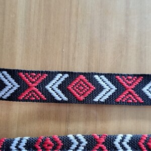 May include: Black woven trim with a repeating pattern of red and white geometric shapes. The trim has a diamond shape in the center of the pattern.