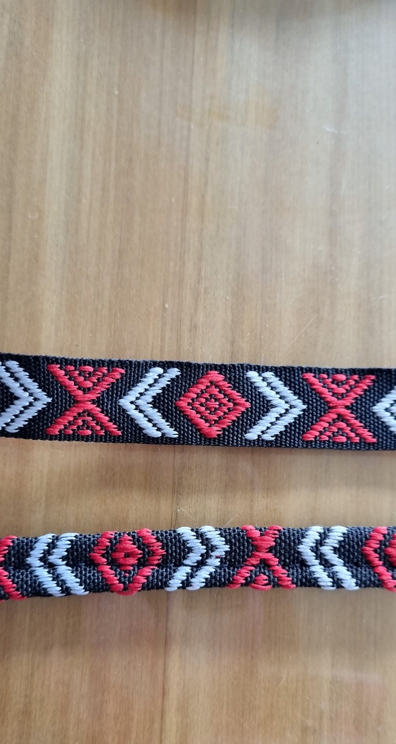 May include: Black woven trim with a repeating pattern of red and white geometric shapes. The trim has a diamond shape in the center of the pattern.