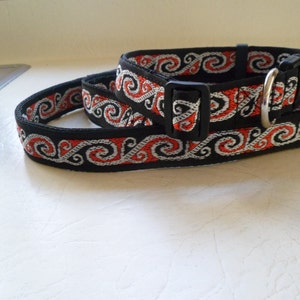May include: Black dog collar with a red and white patterned design. The collar has a silver buckle and a black plastic buckle.