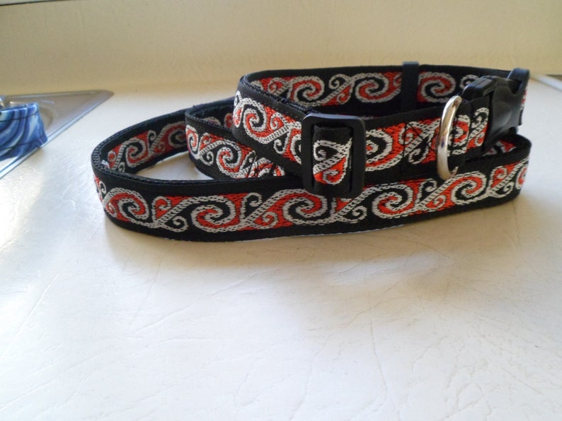 May include: Black dog collar with a red and white patterned design. The collar has a silver buckle and a black plastic buckle.