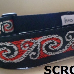 May include: Black dog collar with a red and white patterned design. The pattern features a repeating swirl design. The collar has a metal buckle and a metal D-ring.