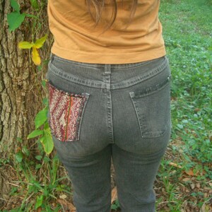 May include: A pair of dark blue jeans with a colorful embroidered patch on the back pocket. The jeans are worn by a person with long brown hair and a brown shirt.