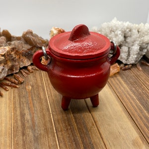May include: A small red cast iron cauldron with a lid and three legs. The cauldron is sitting on a wooden surface.