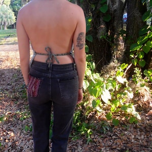 May include: A woman wearing a green and brown halter top with a tied back, dark wash denim jeans, and a small owl tattoo on her arm. She is standing in a wooded area with leaves on the ground.