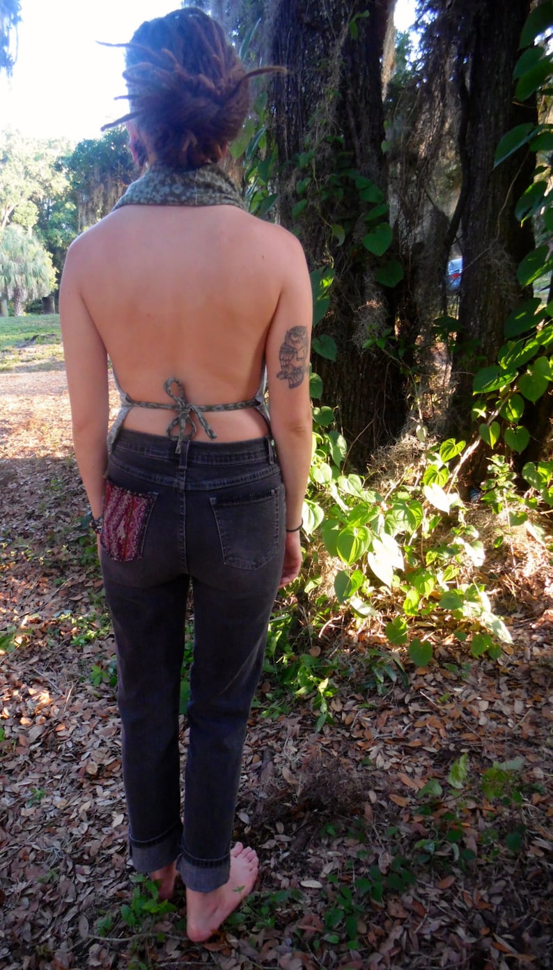 May include: A woman wearing a green and brown halter top with a tied back, dark wash denim jeans, and a small owl tattoo on her arm. She is standing in a wooded area with leaves on the ground.