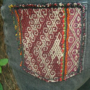 May include: A pair of dark gray denim jeans with a colorful woven patch sewn onto the back pocket. The patch features a geometric pattern in shades of red, white, and green.