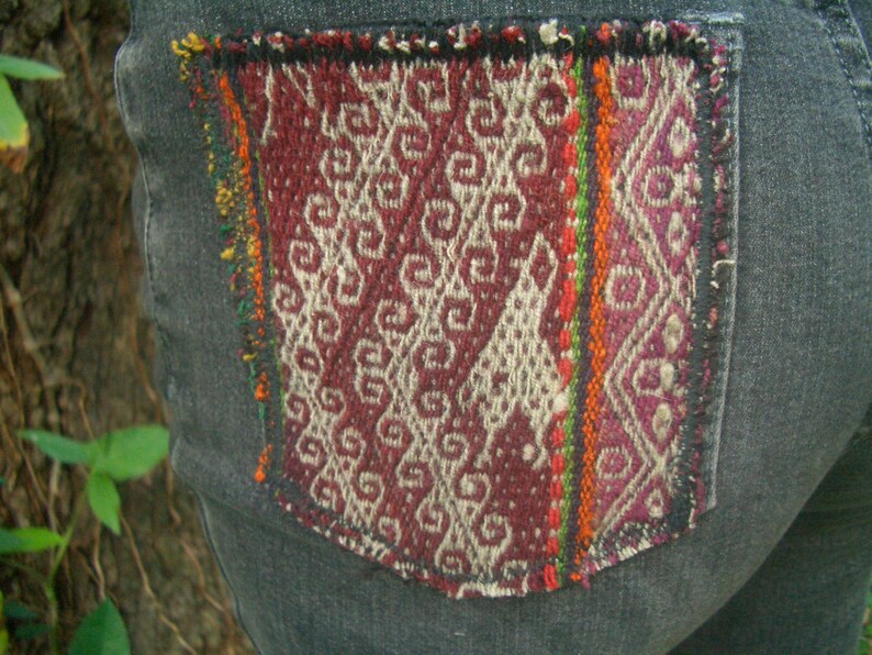 May include: A pair of dark gray denim jeans with a colorful woven patch sewn onto the back pocket. The patch features a geometric pattern in shades of red, white, and green.