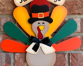 May include: A wooden turkey wall hanging featuring a sign that reads Fall Greetings. The turkey wears a black top hat with an orange band, a black bow tie, and has colourful tail feathers in shades of orange, yellow, and green. The turkey has a tan body.