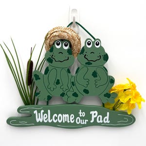 May include: A green wooden sign featuring two cartoon frogs with straw hats. The sign reads "Welcome to our Pad" in white lettering. Decorative yellow flowers and green grass accents the sign.