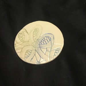 May include: A circular, cream-coloured ceramic coaster with a hand-painted floral design in blue and green hues. The coaster showcases a delicate, stylised flower pattern, adding an artistic touch to any table setting.
