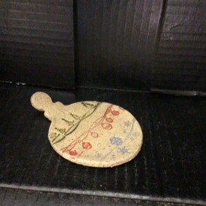 May include: A small, round, ceramic ornament with a loop handle. The ornament is decorated with a winter scene, including trees, snowflakes, and a string of lights.