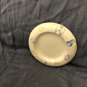 May include: A white ceramic coaster with a floral design in shades of blue, yellow, and green. The coaster has a raised rim and a smooth surface.