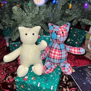 May include: Two plush cat toys, one is white and the other is red and blue plaid, sitting on a green and white gift wrapped box with a Christmas theme.