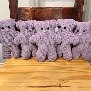 May include: Five purple knitted teddy bears with button eyes and pink noses are lined up on a wooden surface.