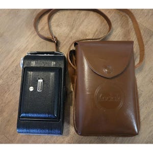 May include: A vintage black Kodak camera with a brown leather case and strap. The camera has a rectangular shape with a silver button and the Kodak logo. The leather case has a flap closure and the Kodak logo embossed on the front.