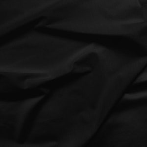 May include: Close-up of black fabric showing folds and shadows. The material has a smooth texture, possibly satin or silk, with a slight sheen. The fabric appears soft and flowing, creating a sense of depth.