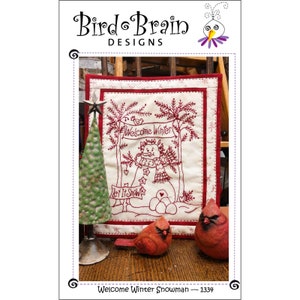 May include: A red and white embroidered quilt block with a snowman and the text "Welcome Winter" and "Let It Snow". Two red cardinal figurines are in the foreground.
