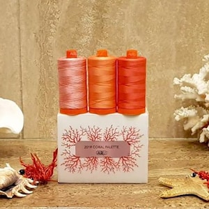 May include: A display of three spools of thread in coral shades, atop a white box with coral-themed artwork and text. The scene is decorated with seashells, coral, and a starfish, creating a coastal theme.