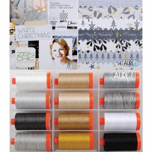 May include: A collection of twelve spools of Aurifil 100% cotton thread in a clear plastic organiser box. The spools feature various shades of white, grey, gold, and black. The background displays a White Christmas theme with patterned paper.