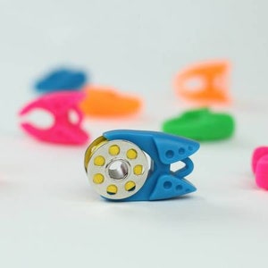 May include: A blue sewing thread spool holder with a gold spool and yellow thread. Other colorful holders in pink, orange, green, and blue are blurred in the background. The holder has a unique design with a metal center.