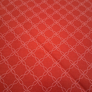 以下が含まれることがあります： 赤色の生地のクローズアップ。白いステッチのパターンが繰り返されています。パターンは、相互接続された様式化された花の形で構成され、テクスチャのある幾何学模様を作成します。生地は柔らかく、マットな仕上がりです。