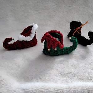 May include: Three crocheted elf shoes in red, green and black. The red shoe has a white fur trim. The green shoe has gold jingle bells. The black shoe has an orange ribbon.