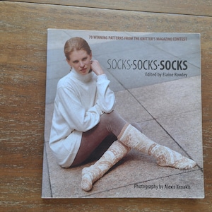 May include: A softcover book titled Socks Socks Socks, edited by Elaine Rowley, featuring 70 winning patterns from the Knitter's Magazine contest. The cover displays a person wearing a white turtleneck sweater and patterned knit socks.