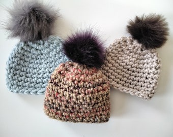 May include: Three crocheted hats with faux fur pom-poms. The hats are in light blue, beige, and a multi-colored pattern.