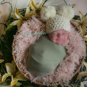Puede incluir: Un bebé recién nacido con un gorro blanco de ganchillo con pompones, envuelto en una manta verde, y acostado en una cesta de mimbre forrada con piel sintética rosa.