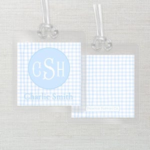 May include: Clear plastic luggage tag with a light blue gingham pattern. The tag features a light blue circle with the initials "CSH" in white, and the name "Charlie Smith" below. The second tag has the text "PRINTED POPPIES CO."