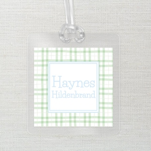 May include: Clear luggage tag with a green and white plaid pattern. The tag features a light blue border and the names "Haynes Hildenbrand" in light blue. A clear plastic loop is attached at the top.