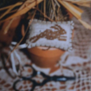 May include: A white fabric pillow with a brown embroidered bunny design. The pillow is sitting on a brown pot with a straw bow on top. The pot is sitting on a white crocheted surface.