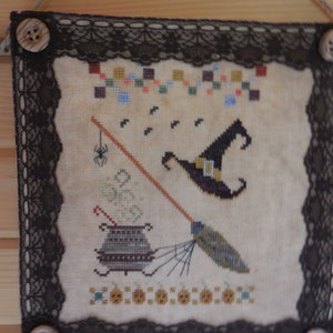 May include: A beige cross-stitch wall hanging with a black lace border and wooden buttons. The design features a witch's hat, a cauldron, a broom, and pumpkins.