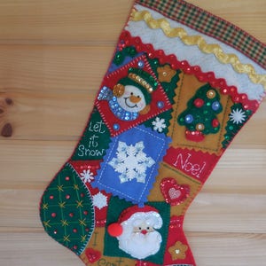 Puede incluir: Una media de Navidad roja, verde y dorada con una variedad de aplicaciones de fieltro, incluyendo un muñeco de nieve, un árbol de Navidad, un copo de nieve y las palabras "Let it Snow", "Noel" e "I Santa".
