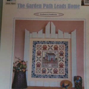 Könnte beinhalten: Ein Zählkreuzstich-Muster für eine Quiltdecke mit einem Häuschen, einem Lattenzaun und einem Gartenweg. Das Muster trägt den Titel "The Garden Path Leads Home" und ist Teil der Serie "Garden Gate Quilt III". Das Muster wurde von Linda Myers entworfen.