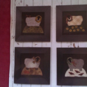 May include: Four framed pictures of wool appliqué sheep, each with a different design. The frames are black and the background is white wood. The text "Yearly Ewe" and "Four 8" x 8" Wall Appliqué Pictures" is written below the frames.