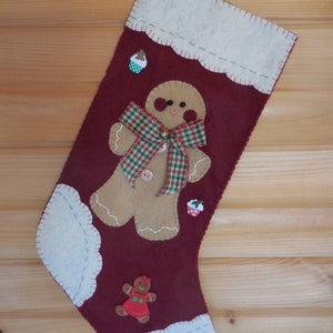 May include: Red felt Christmas stocking with a gingerbread person appliqué. The gingerbread person has a plaid bow, a button nose, and a bell. The stocking has white felt trim and a white felt cuff.